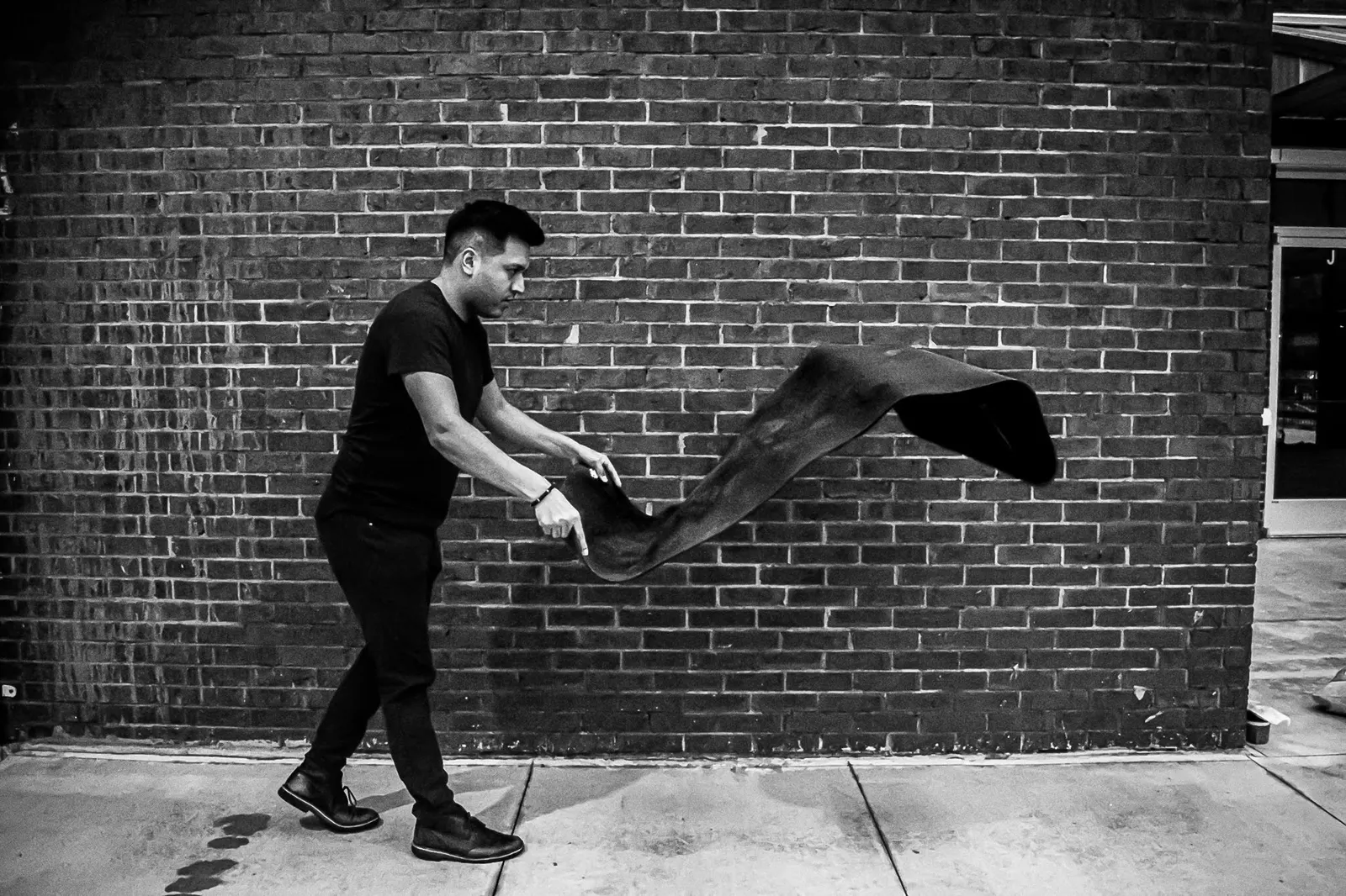 Black and white photo of a man swinging a yoga mat through the air in front of a brick wall